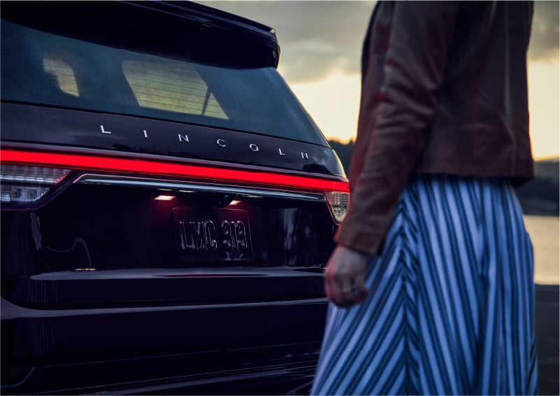 A person is shown near the rear of a 2023 Lincoln Aviator® SUV as the Lincoln Embrace illuminates the rear lights | Holmes Tuttle Lincoln in Tucson AZ